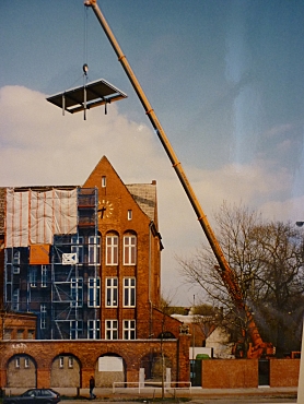 Auf dem Bild ist ein Kran zu sehen, der ein Bauteil zur Schule im Hintergrund hochhebt.